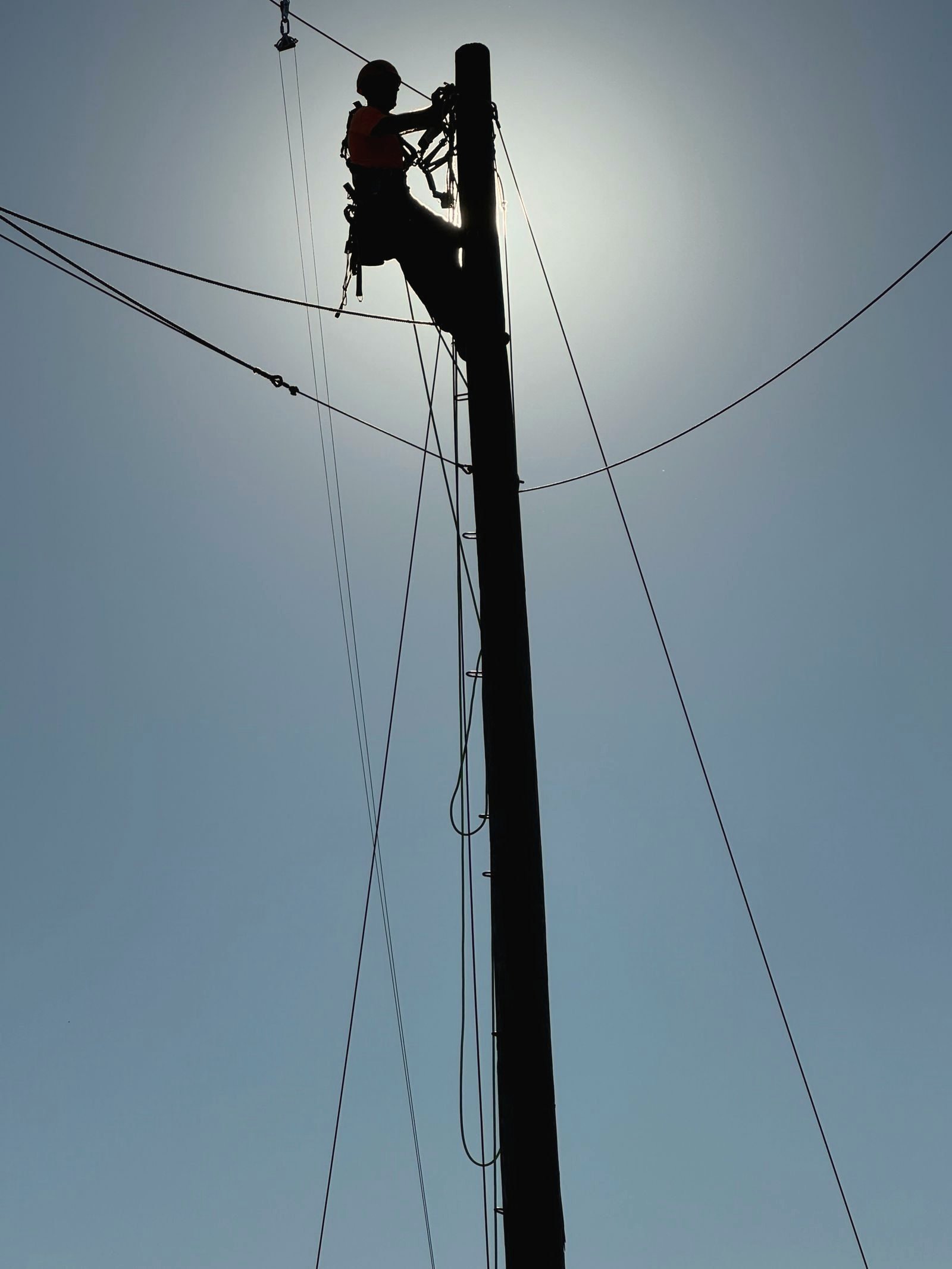 Lineman in hard hat and harness silhouetted at the top of a wooden pole
